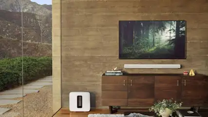 Innenraum mit Fernseher, Lautsprecher, Kommode und weißem Subwoofer vor einer Wand. Draußen ist eine Berglandschaft sichtbar.