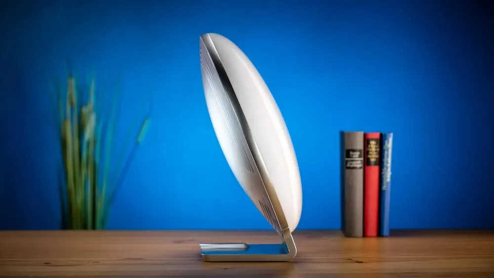 Eine weiße Tageslichtlampe steht seitlich auf einem Holzschreibtisch vor einem blauen Hintergrund mit Büchern und Gras.