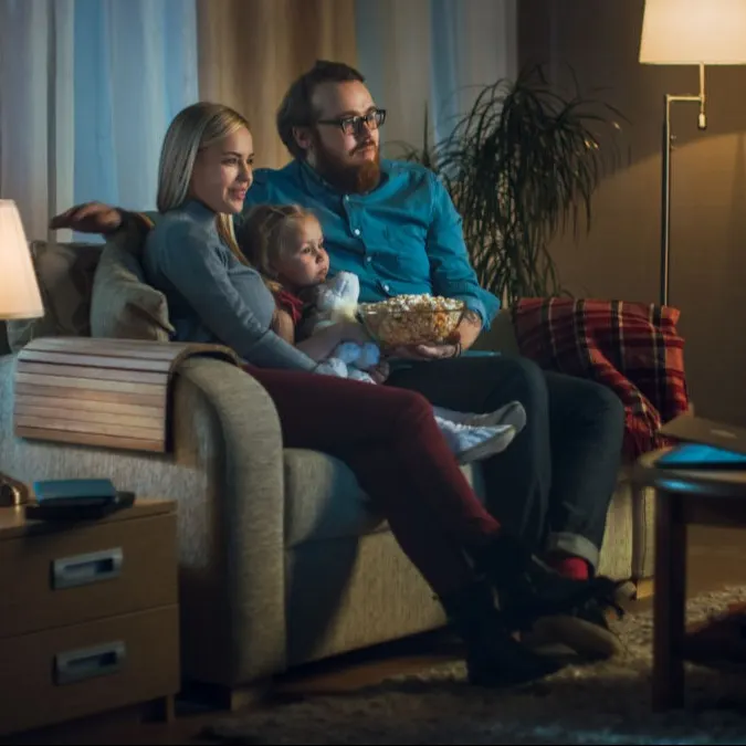 Eine Familie sitzt auf einem Sofa und schaut fern, der Mann hält eine Schüssel Popcorn. Ein Tisch und Regale sind im Hintergrund.