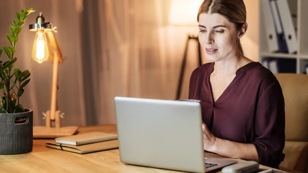 Eine Frau sitzt an einem Schreibtisch mit Laptop, Lampe, Pflanze und Notizbüchern.