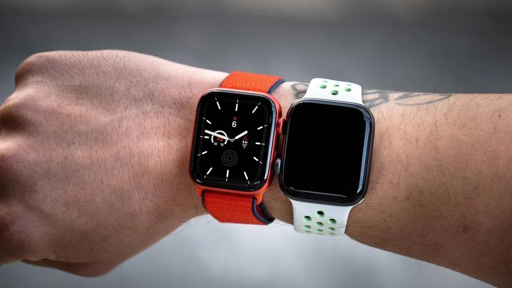 Zwei Smartwatches am Handgelenk. Eine hat ein oranges Armband und ein Zifferblatt, die andere ist schwarz mit einem weißen Armband.