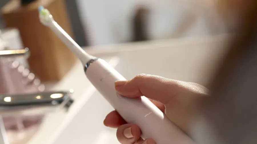 Eine Hand hält eine weiße elektrische Zahnbürste mit einem farbigen Bürstenkopf. Im Hintergrund sind ein Waschbecken und Badezimmerartikel.