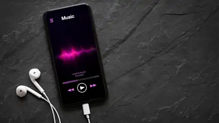 Ein Smartphone liegt neben weißen Kopfhörern, die an das Smartphone angeschlossen sind. Man sieht einen Musik-Player auf dem Bildschirm.