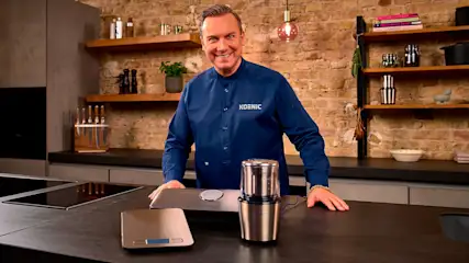In einer Küche steht ein Mann in blauem Hemd hinter einer Küchenwaage und einer Kaffeemühle auf einer dunklen Arbeitsplatte.