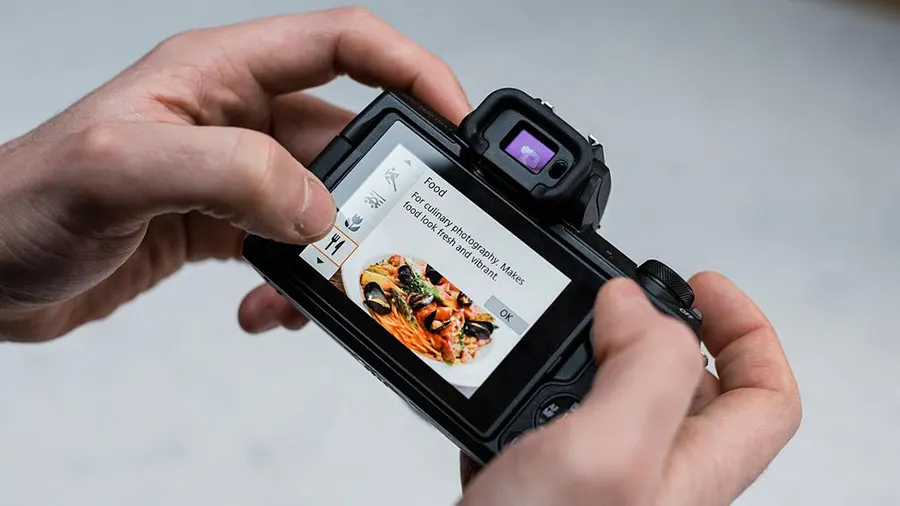 Eine schwarze Kamera mit einem Touchscreen, auf dem ein Teller Pasta angezeigt wird, wird von zwei Händen gehalten.