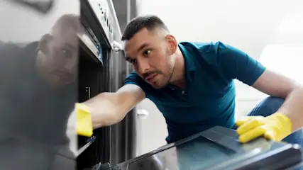 Ein Mann mit gelben Handschuhen reinigt einen schwarzen Ofen mit einem Lappen. Er trägt ein blaues Poloshirt und Jeans.