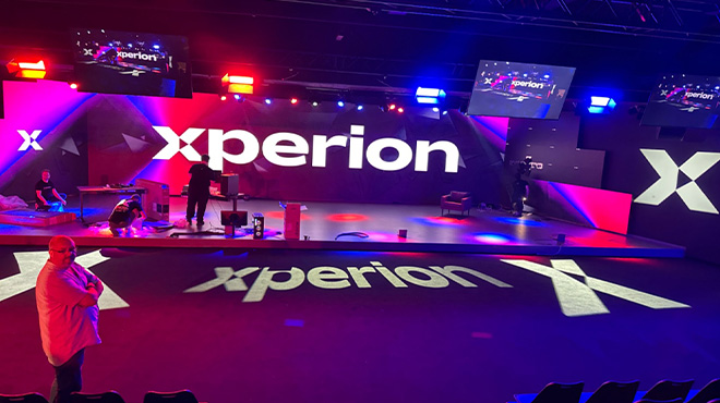 Eine Bühne mit dem Schriftzug "xperion" in Weiß, beleuchtet in Rot und Blau. Personen stehen auf der Bühne.