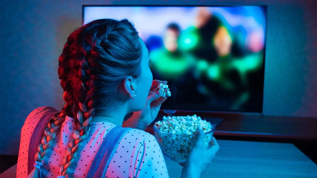 Frau mit Zöpfen sitzt vor dem Fernseher und isst Popcorn aus einer Glasschüssel bei blauem Licht.