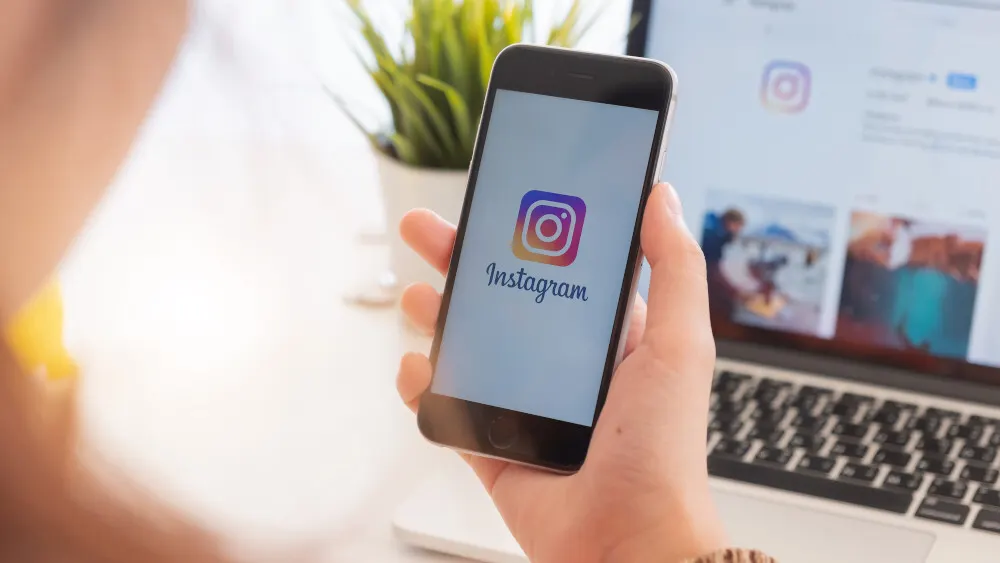 Hand hält ein Smartphone mit dem Instagram-Logo vor einem Laptop. Eine Pflanze steht im Hintergrund.