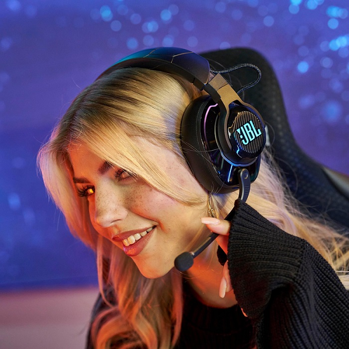 Eine Frau lächelt mit einem JBL-Headset und einem Mikrofon vor einem blauen Hintergrund. Sie trägt einen schwarzen Pullover.