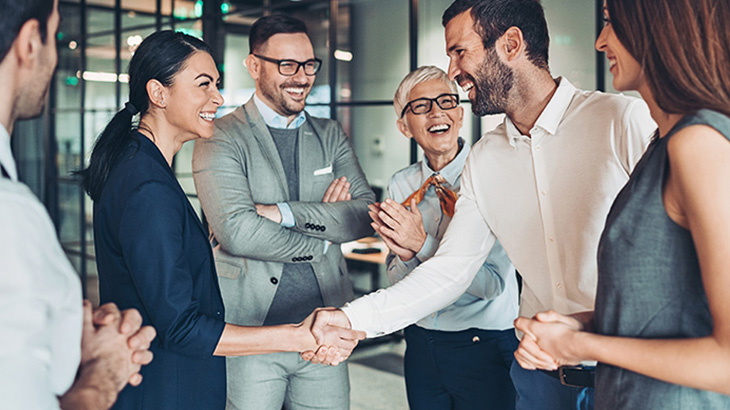 Mehrere Personen in formeller Kleidung stehen zusammen. Einige schütteln sich die Hände und lachen. Die Szene wirkt geschäftlich und freundlich.