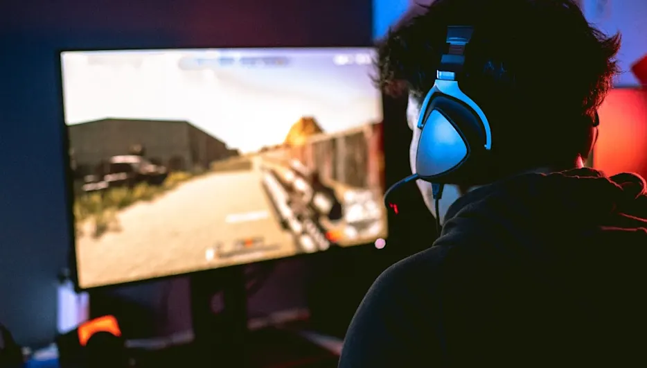 Person mit Kopfhörern vor einem Computerbildschirm, der eine Wüstenlandschaft in einem Videospiel zeigt.