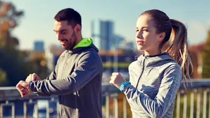 Zwei Personen in Sportkleidung stehen auf einer Brücke und schauen auf ihre Uhren. Im Hintergrund sind Bäume und Gebäude.