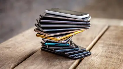 Ein Stapel von etwa zwölf Mobiltelefonen unterschiedlicher Farben liegt auf einem Holztisch.