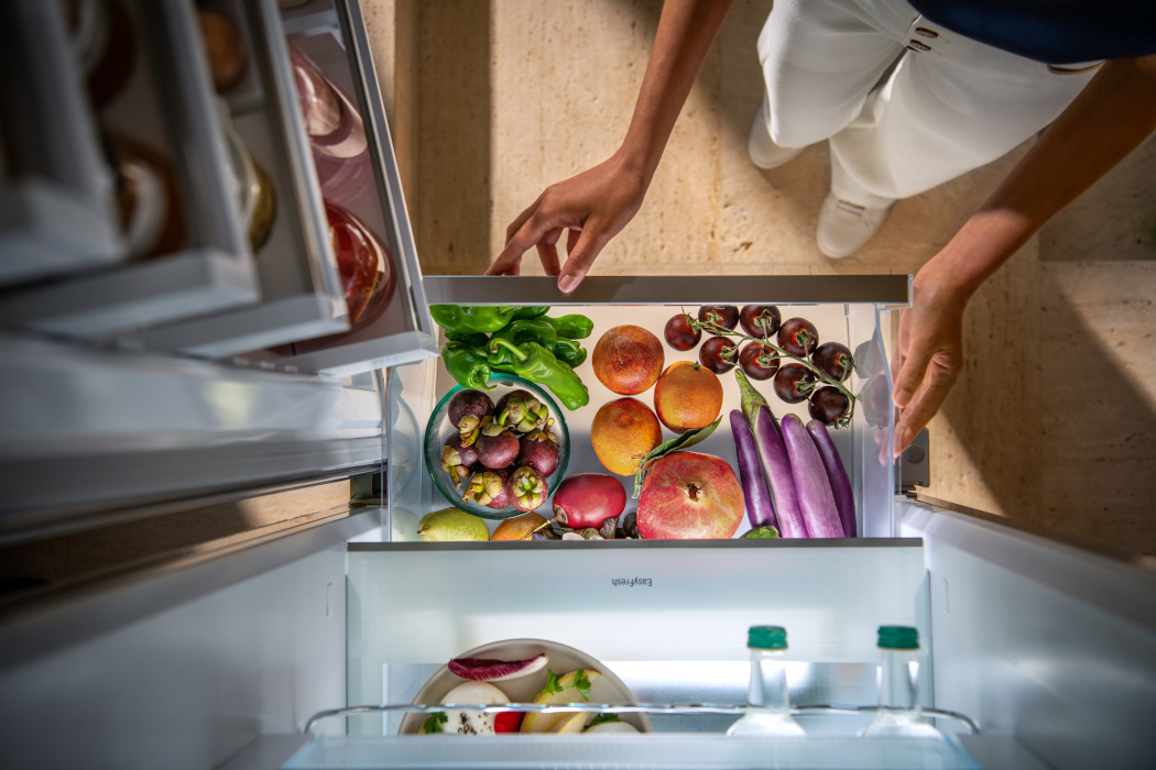Eine Person öffnet eine Kühlschublade mit Obst und Gemüse wie Mangos, Auberginen und Paprika.
