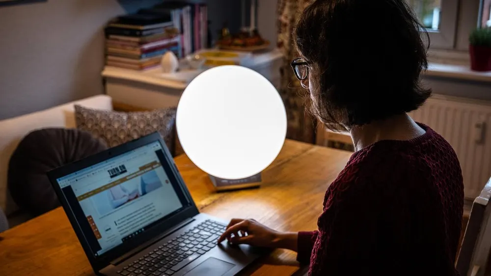 Frau mit Brille arbeitet am Laptop auf einem Holztisch. Eine runde Lampe steht daneben. Im Hintergrund sind Bücher und ein Sofa.