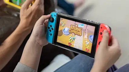 Eine Person hält eine Nintendo Switch mit blauem und rotem Controller, auf dem "Animal Crossing" läuft. Andere Person mit gelbem Gerät im Hintergrund.