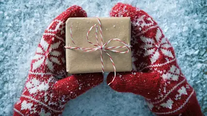 Gerahmtes, braunes Geschenk mit rot-weißem Band in rot-weißen Handschuhen auf Schnee.