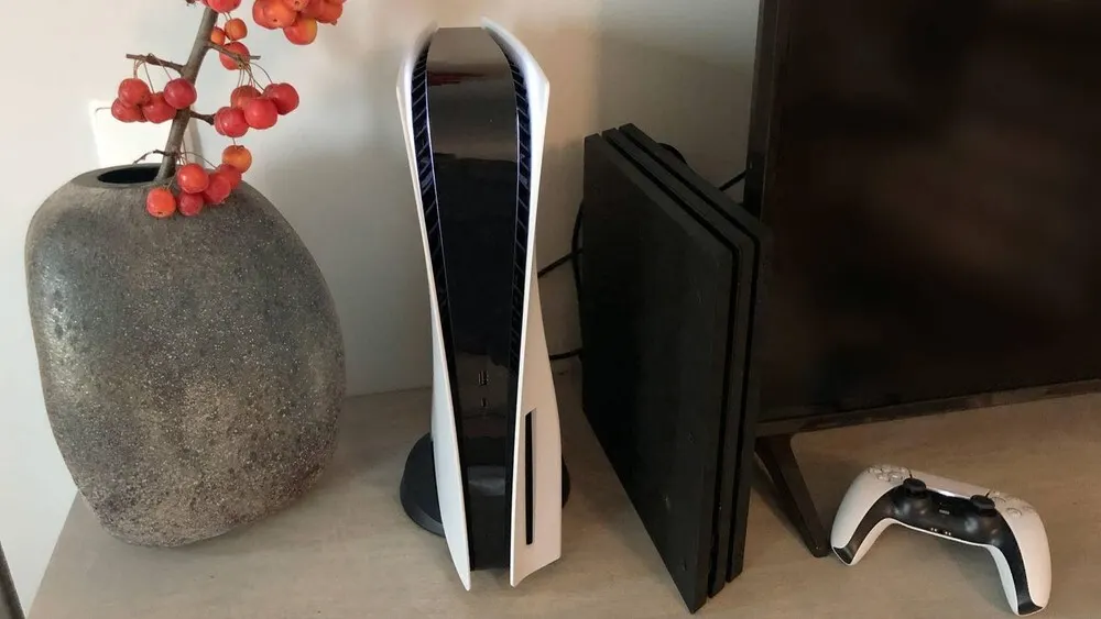 Auf einer hellen Fläche stehen eine graue Vase mit roten Beeren, eine weiße Spielkonsole, eine schwarze Konsole, ein Fernseher und ein Controller.