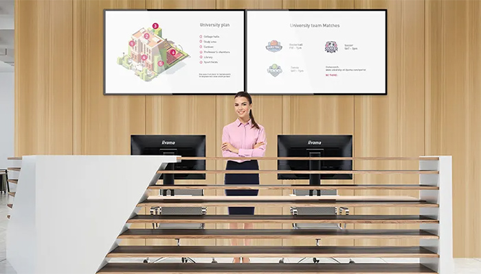 Eine Frau steht hinter einer weißen Rezeption mit zwei Monitoren. Über ihr hängen zwei große Bildschirme an einer Holzwand.