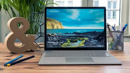 Ein Laptop mit blauem Hintergrundbild steht auf einem Holztisch, neben einem "&"-Zeichen, Stiften und einem Stifthalter.