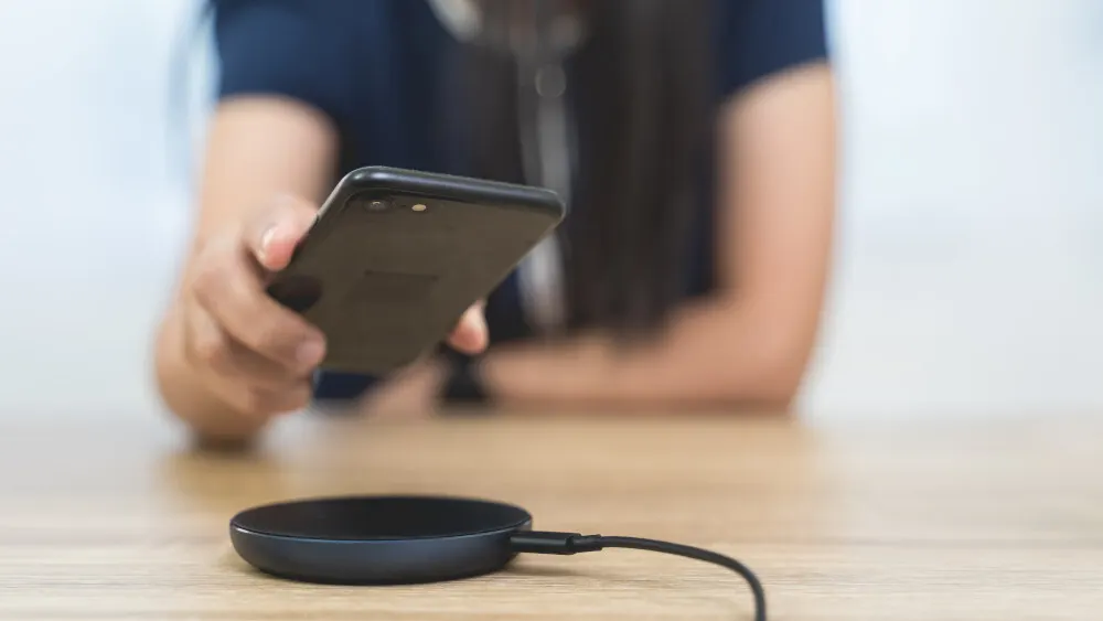 Eine Person hält ein schwarzes Handy über ein drahtloses Ladegerät auf einem Holztisch.