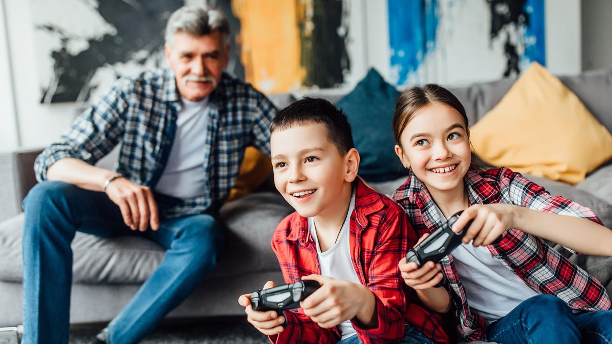 Zwei Kinder und ein älterer Mann sitzen vor und auf einer Couch und spielen mit Controllern.