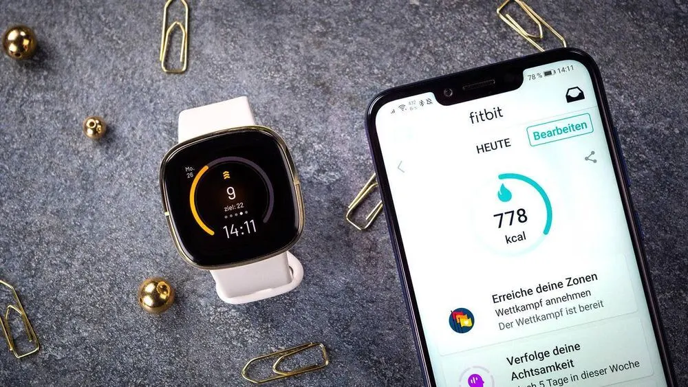 Eine Smartwatch mit weißem Armband und ein Smartphone mit Fitbit-App liegen auf grauem Untergrund, umgeben von Büroklammern.