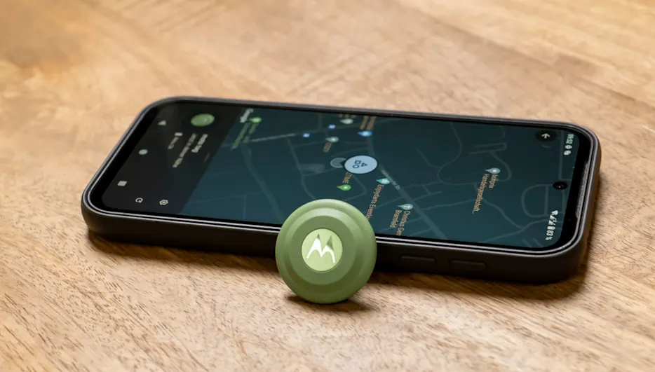 Ein Smartphone mit Karten-App liegt neben einem grünen, runden Motorola-Tracker auf einem Holztisch.