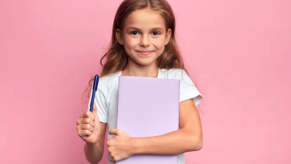 Ein Mädchen hält einen lila Ordner und einen blauen Stift vor einem rosafarbenen Hintergrund. Sie trägt ein weißes T-Shirt.