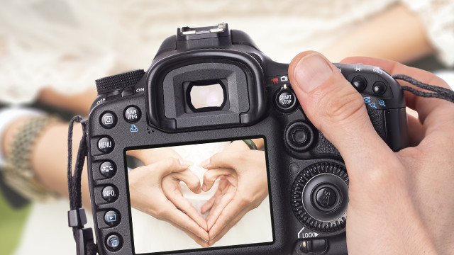 Eine schwarze Kamera in der Hand zeigt ein Herz, das mit Händen auf dem Bildschirm geformt wird.