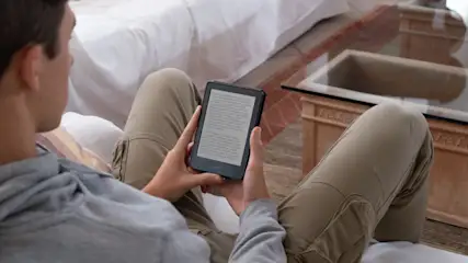 Eine Person sitzt und hält ein schwarzes Lesegerät mit Text auf dem Bildschirm in den Händen; ein Tisch steht im Hintergrund.