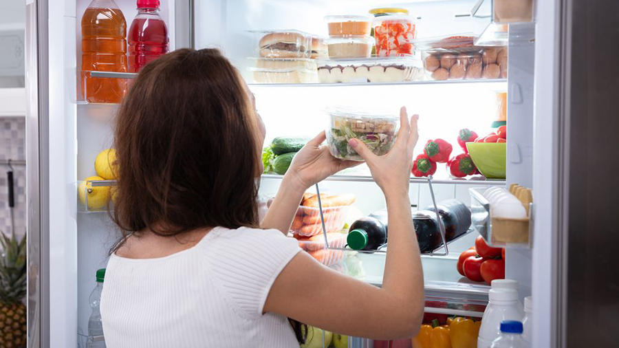 Eine Frau in Weiß nimmt einen Behälter mit Salat aus einem vollen Kühlschrank. Flaschen, Obst und Gemüse sind sichtbar.