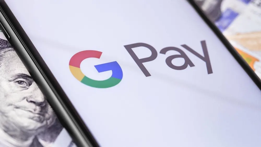 Auf dem Bildschirm eines schwarzen Mobiltelefons befindet sich das Logo von Google Pay. Im Hintergrund sind US-Dollar-Scheine zu sehen.