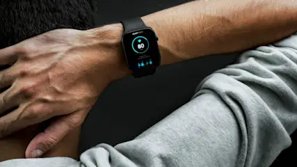 Eine Person trägt eine schwarze Smartwatch, die eine Herzfrequenz von 80 Schlägen pro Minute anzeigt. Die Person trägt ein graues Sweatshirt.