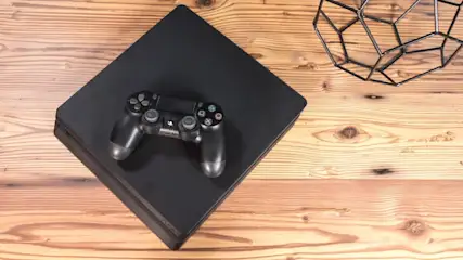 Ein Spielcontroller liegt auf einer schwarzen Spielkonsole. Diese befindet sich auf einem Holztisch. Rechts ist eine schwarze Deko-Drahtkugel.