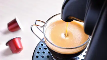 Eine Kaffeemaschine füllt Kaffee in ein Glas. Zwei rote Kaffeekapseln liegen daneben auf einer weißen Oberfläche.