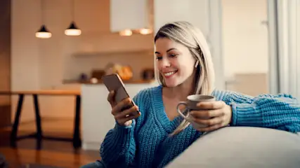 Eine Frau im blauen Pullover hält ein Handy und eine Tasse in einem Wohnzimmer mit Lampen.