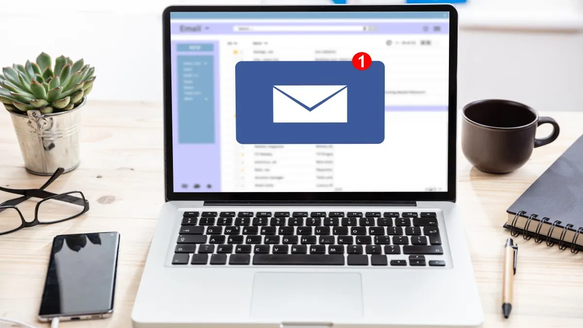 Ein Laptop zeigt ein E-Mail-Symbol mit einer roten "1". Daneben stehen ein Notizbuch, ein Stift und eine Tasse; links eine Pflanze und eine Brille.