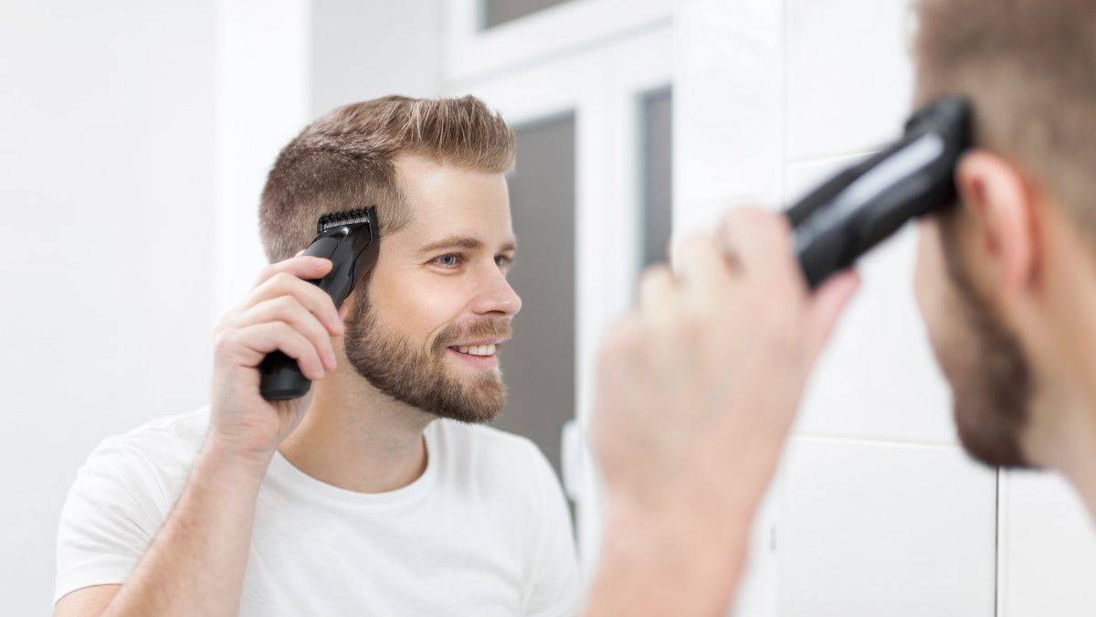 Ein junger Mann schneidet sich mit einer Haarschneidemaschine vor einem Spiegel die Haare.