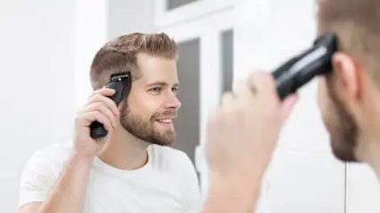 Ein junger Mann schneidet sich mit einer Haarschneidemaschine vor einem Spiegel die Haare.