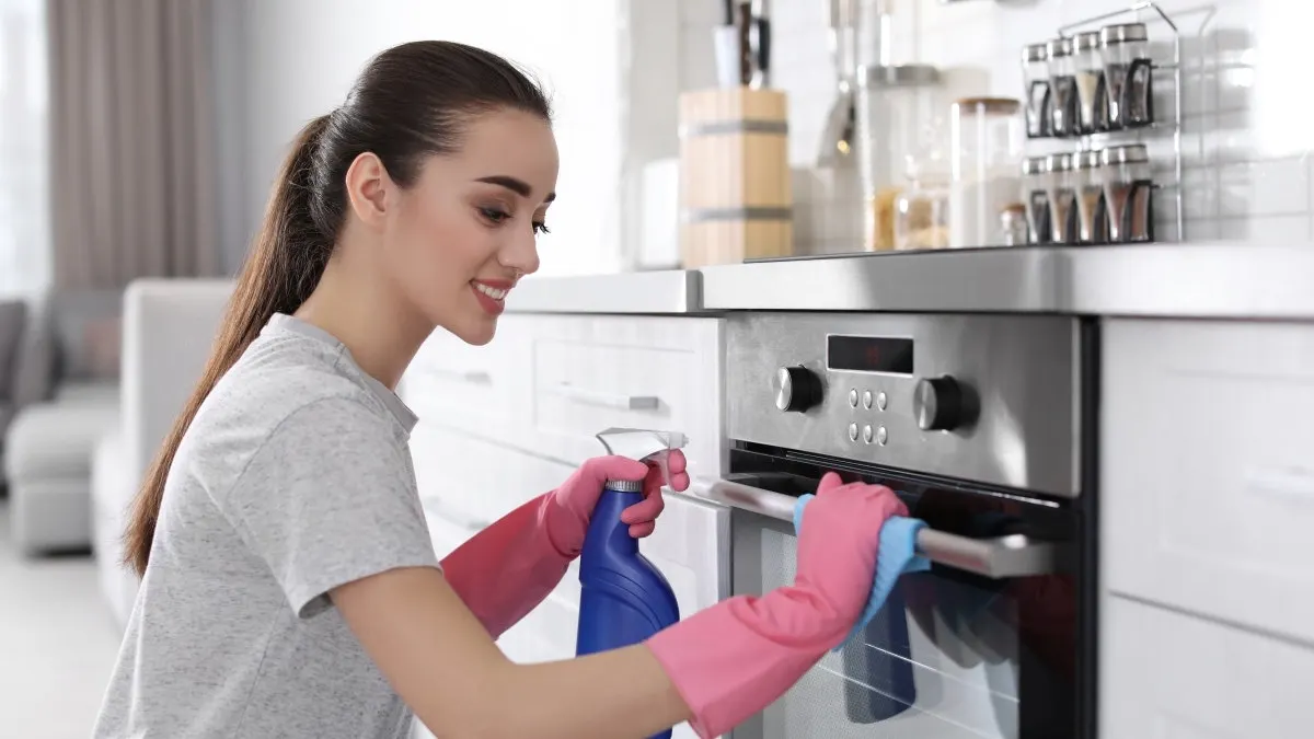 Eine Frau mit rosa Handschuhen putzt einen silbernen Ofen mit einem blauen Spray in einer Küche.
