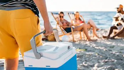 Person trägt Kühlbox am Strand. Strandkörbe und Meer im Hintergrund.