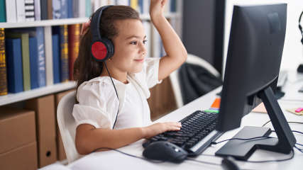 Junges Mädchen mit Headset sitzt vor einem Computerbildschirm an einer ergonomischen Tastatur und meldet sich