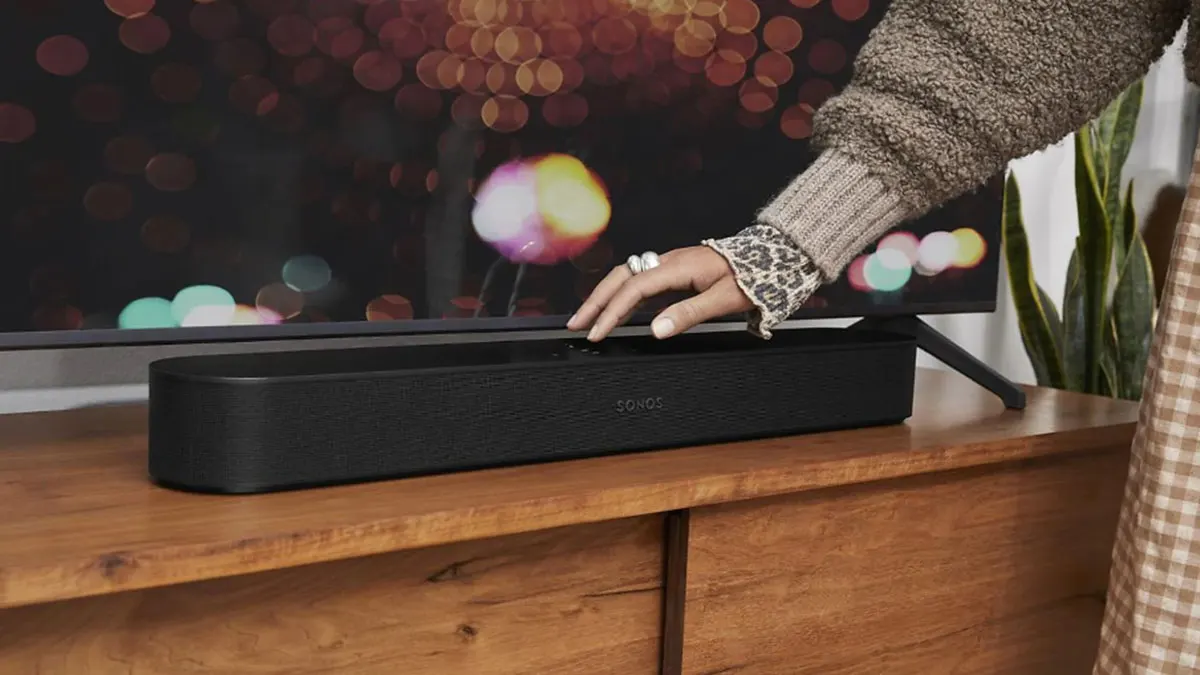 Sonos Soundbar auf einer Kommode vor einem Fernseher. Eine Hand bedient die Soundbar.