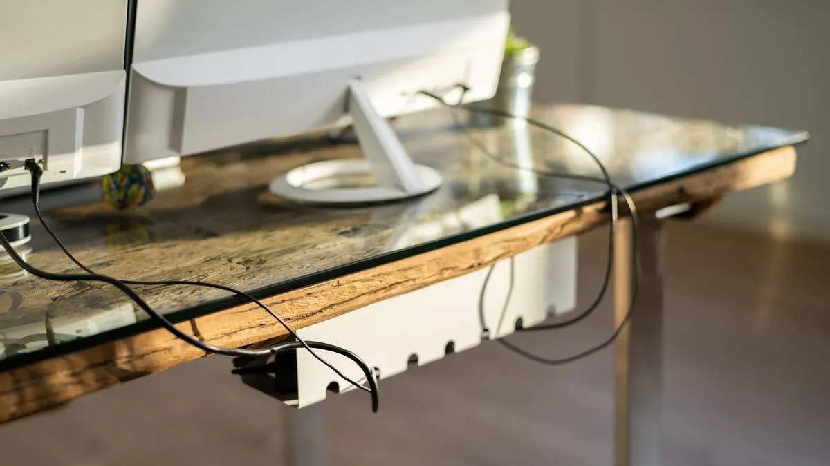 Ein Schreibtisch mit Glasplatte und Holzrahmen steht im Raum. Ein weißer Computermonitor und Kabel sind sichtbar.