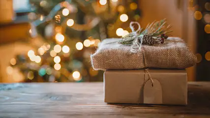 Zwei Geschenke, eines in Jute gehüllt, mit Tannenzweig und Zapfen, auf einem Holztisch vor einem Weihnachtsbaum mit Lichtern.