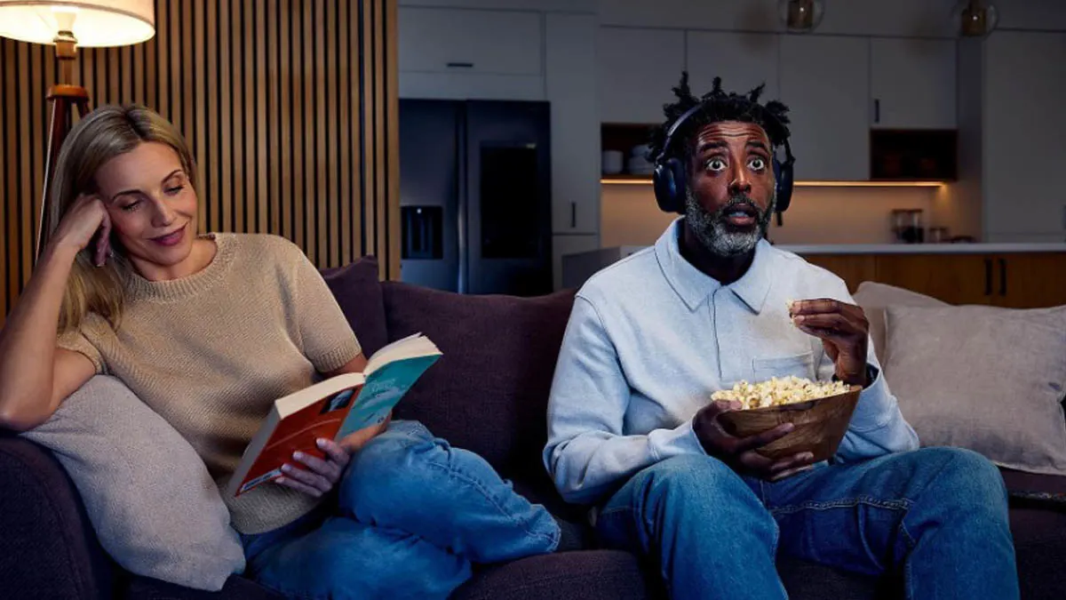 Eine lesende Frau sitzt neben einem Mann mit Kopfhörern auf der Couch. Der Mann sieht gebannt nach vorne und hält eine Schüssel Popcorn auf dem Schoß.