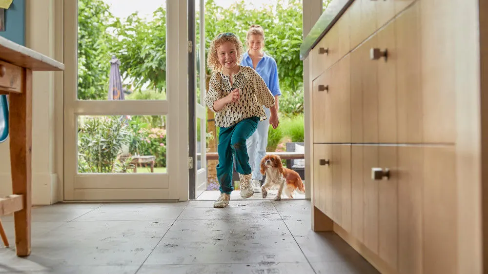 Ein Kind in gemustertem Oberteil und grüner Hose rennt in ein Zimmer, gefolgt von einer Frau und einem Hund. Im Hintergrund ist ein Garten sichtbar.