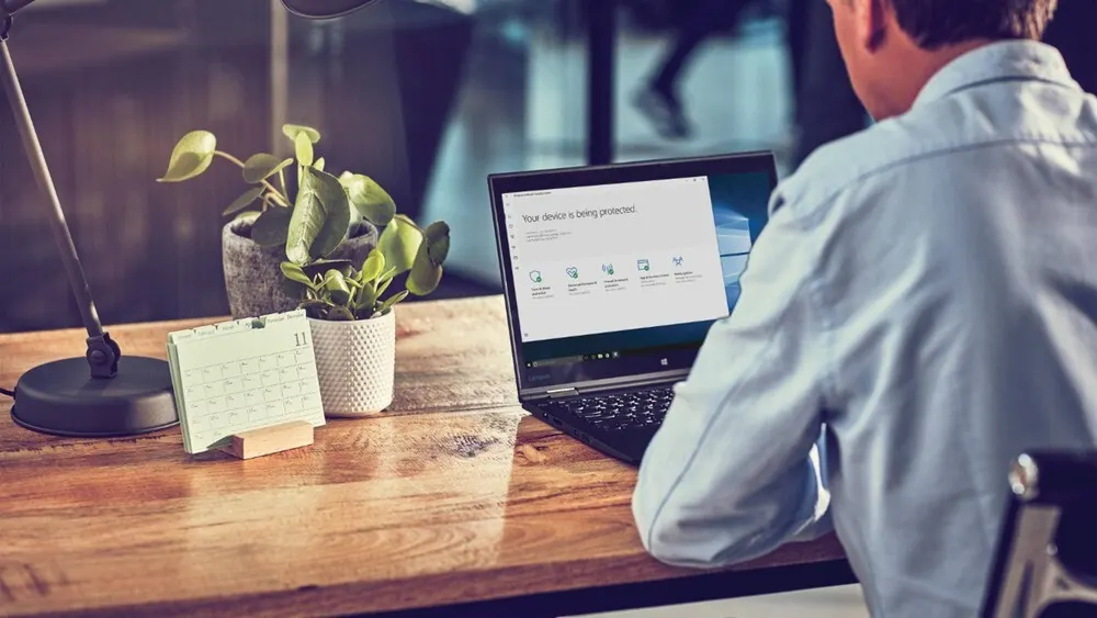 Mann sitzt vor Laptop an Holztisch. Auf dem Tisch stehen eine Lampe, zwei Pflanzen und ein Kalender.